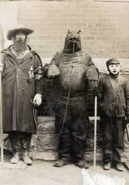 A sepia-toned vintage photograph shows three figures standing against a brick wall: a bearded man in a hat and coat on the…