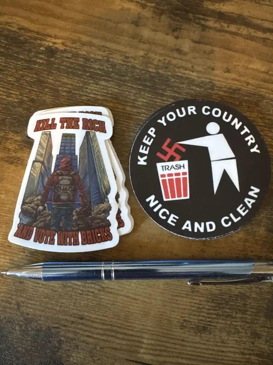 Two stickers sit on a wooden surface next to a pen. One sticker shows a hooded figure with a backpack standing among rubble…