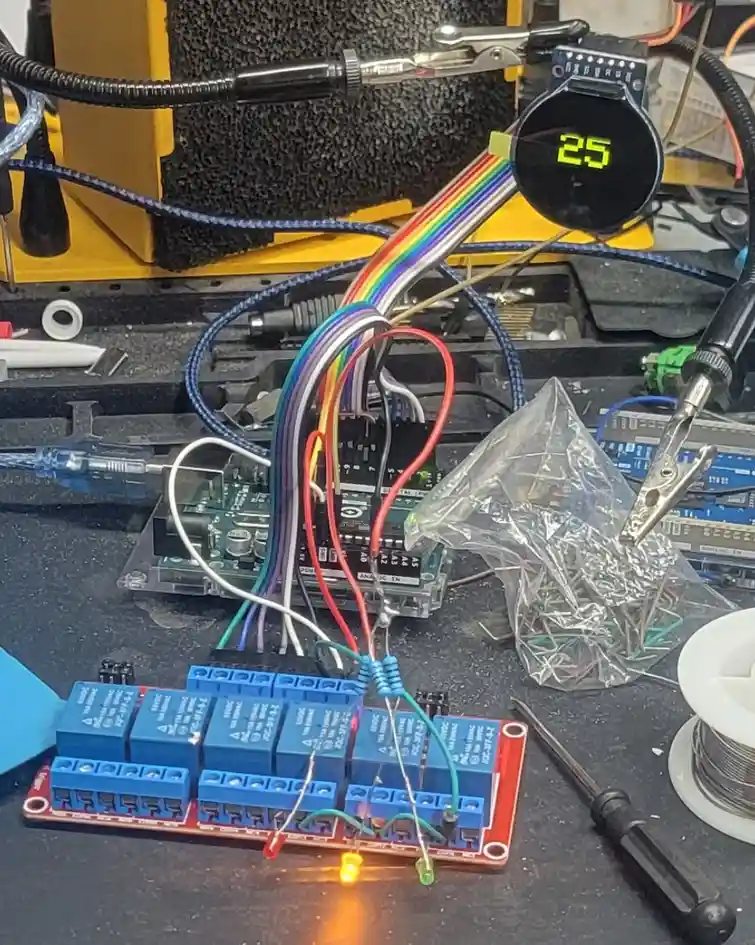 A cluttered electronics workbench shows a digital clock displaying '25' while connected to a microcontroller and relay board…