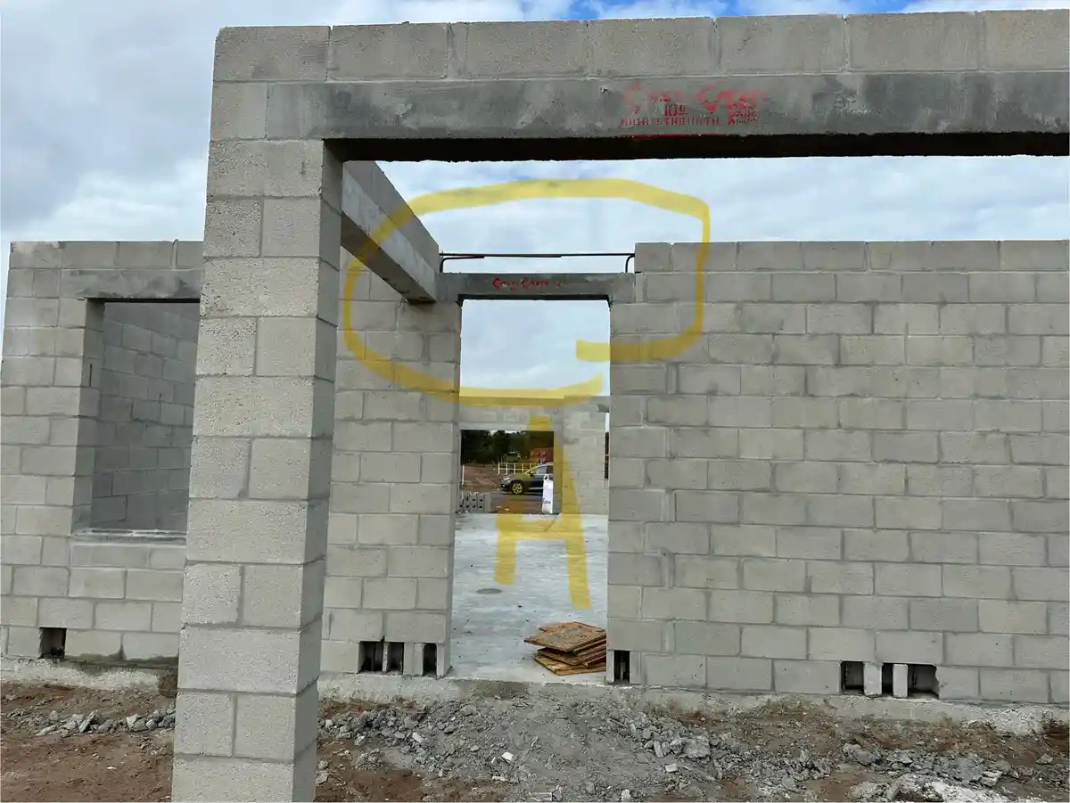 An unfinished concrete block building with a large opening framed by gray blocks under a cloudy sky. A large yellow letter…