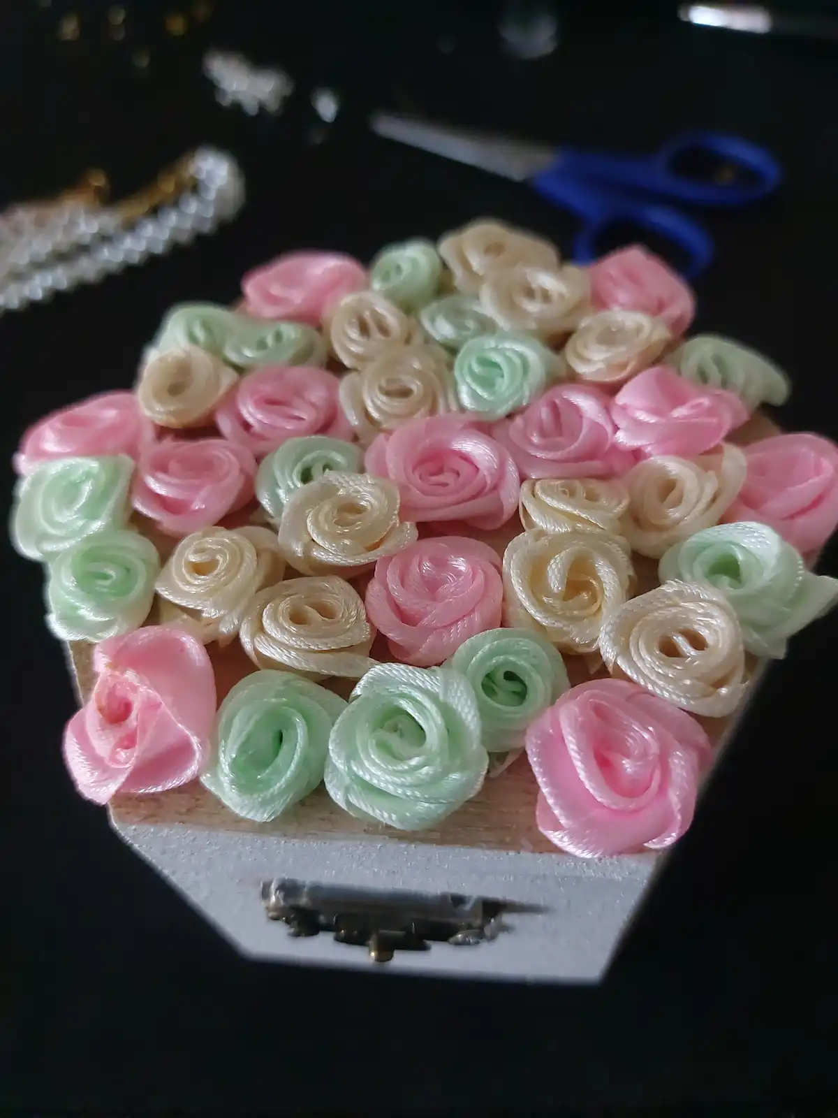 A handmade coffin decorated with pink, green, and cream-colored ribbon roses sits on a dark surface, with crafting tools…