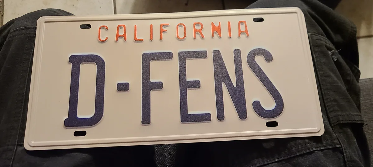 A close-up photo shows a California license plate resting on dark denim jeans. The plate has orange 'CALIFORNIA' text at the…