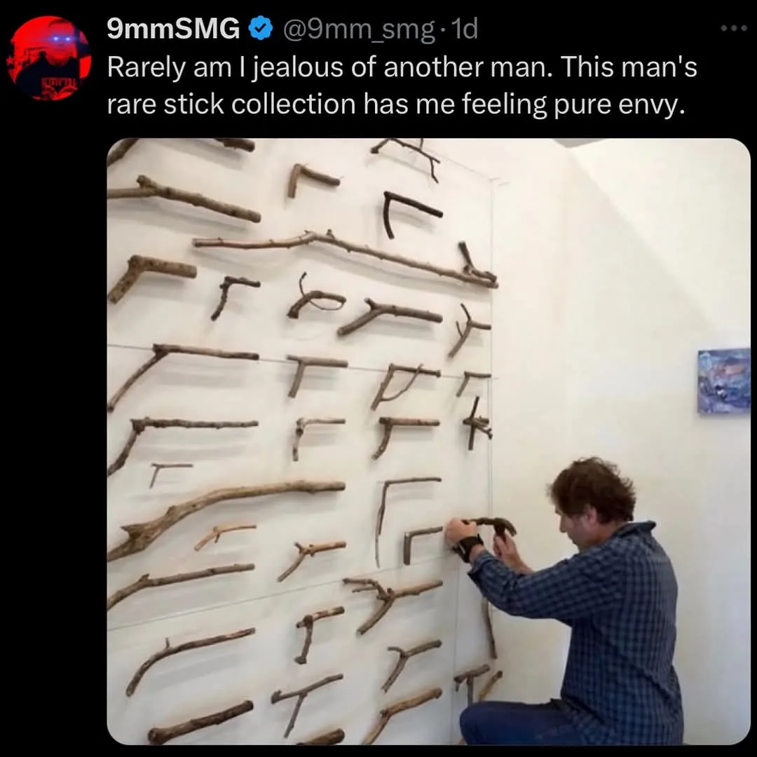 A man in a plaid shirt kneels before a white wall meticulously arranging numerous sticks of various shapes and sizes into an…