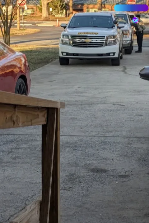 A person in a dark uniform with a badge stands beside a white Chevrolet Suburban in a driveway. A wooden table leg is in the…
