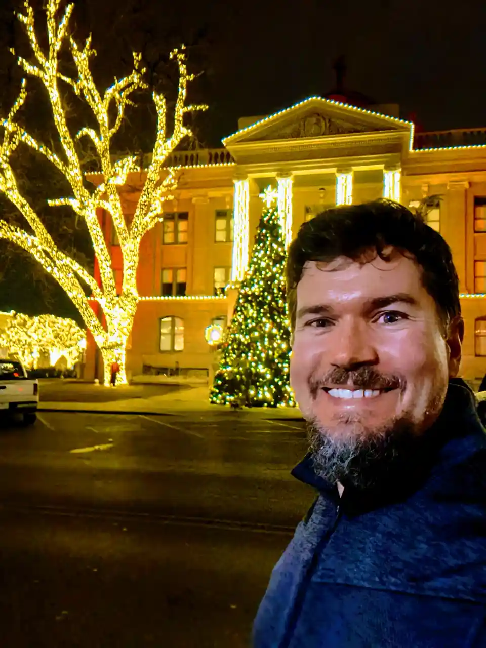 A man with a beard and dark hair smiles at the camera while standing outdoors at night in front of a large, brightly lit…