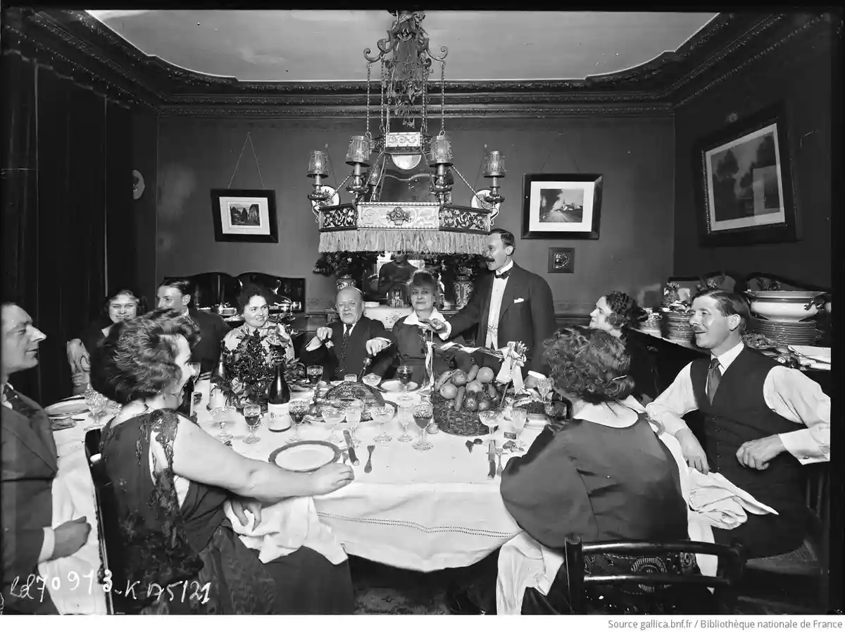 a vintage black-and-white photo shows a group of formally dressed people gathered around a lavishly set dining table with a…