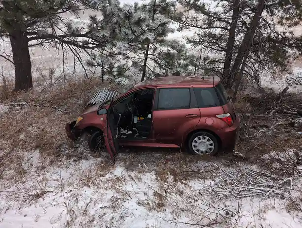A maroon car is wrecked in a snowy, wooded area with its driver's door open and front bumper damaged. Snow covers the ground…