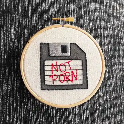 An embroidered image of a floppy disk on fabric inside a wooden embroidery hoop, with the words 'NOT PORN' stitched in red…