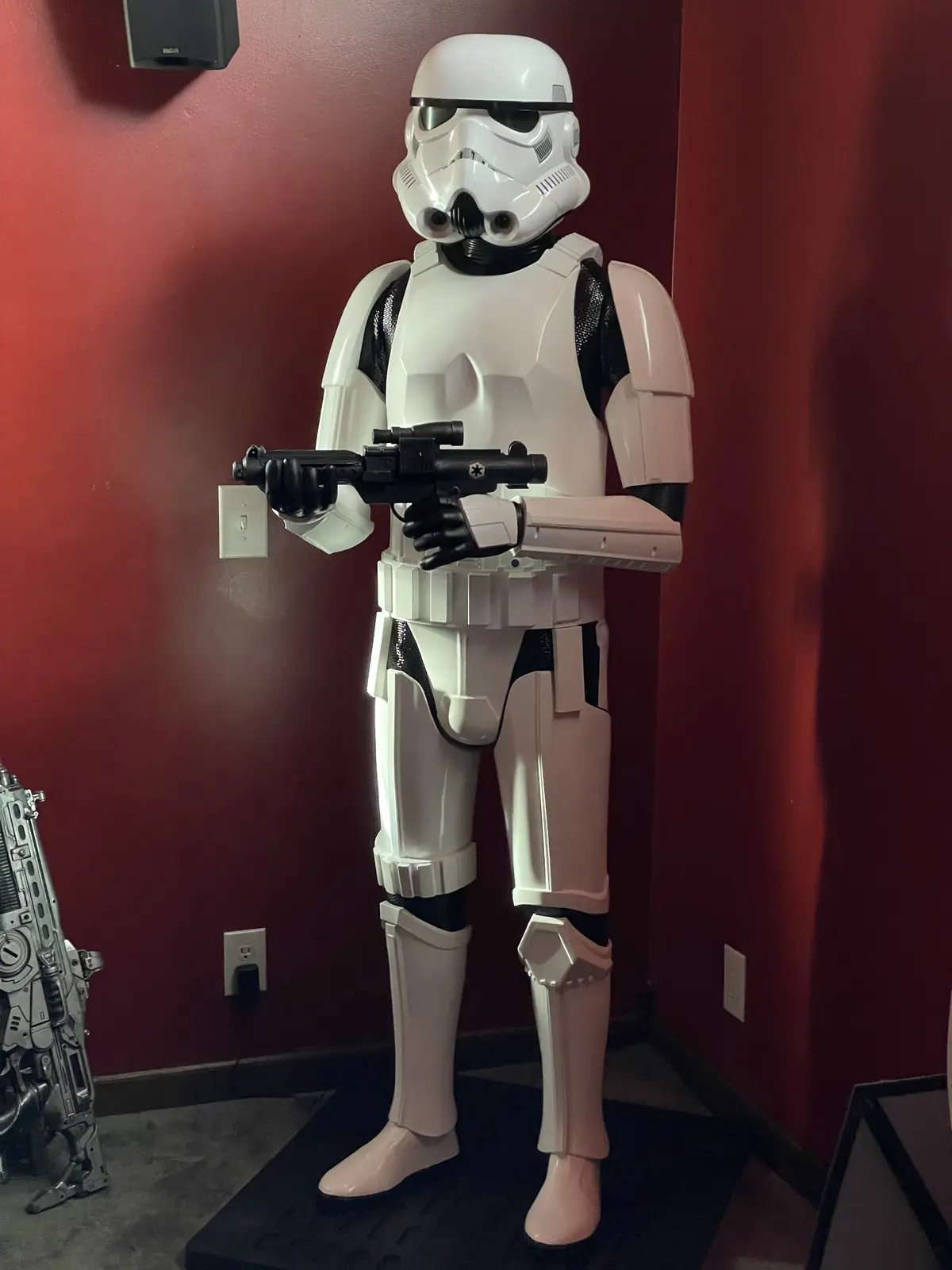 A full-body Stormtrooper costume stands in a room with red walls, holding a blaster rifle. The text overlaid on the image…
