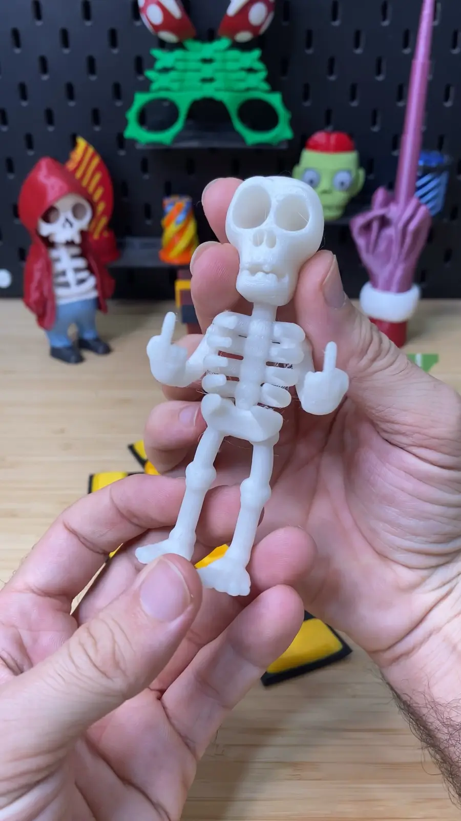 A person holds a small white 3D-printed skeleton figurine that is making a rude gesture with both hands. The background…