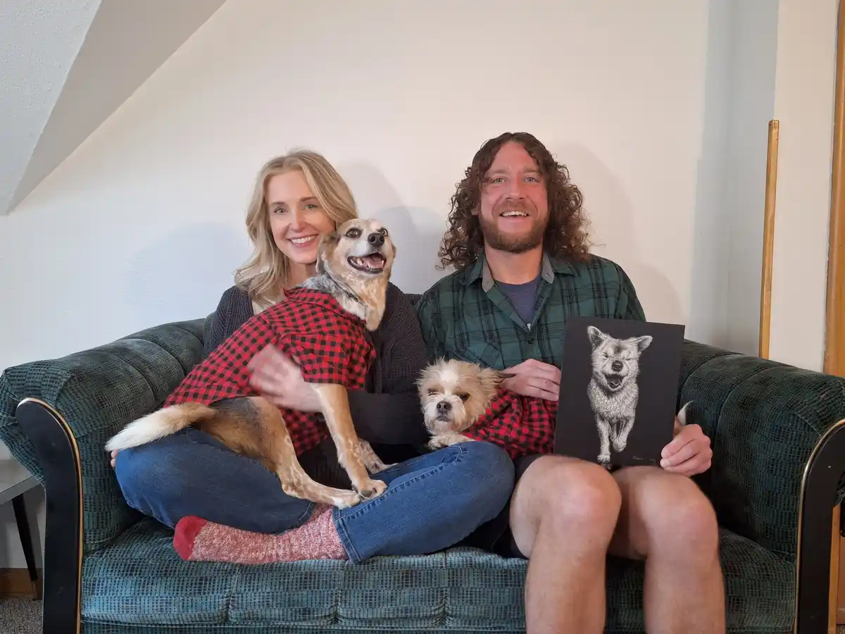 A man and woman sit on a green couch with two dogs; the man holds up a black-and-white drawing of one of the dogs, which is…