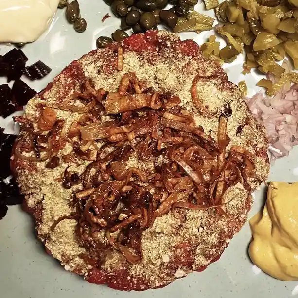 A close-up photo of a raw beef tartare patty on a plate, topped with crispy fried onions and surrounded by capers, pickled…