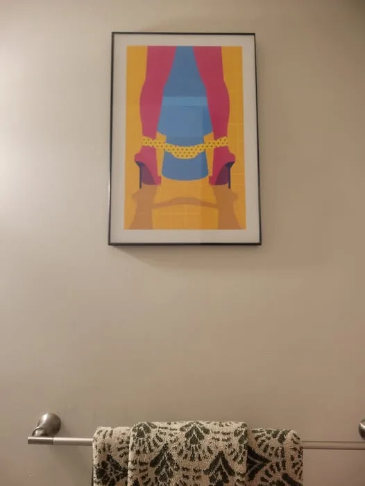 A framed, stylized pop-art print of a woman's legs in pink high heels and a blue skirt hangs on a beige wall above a towel…