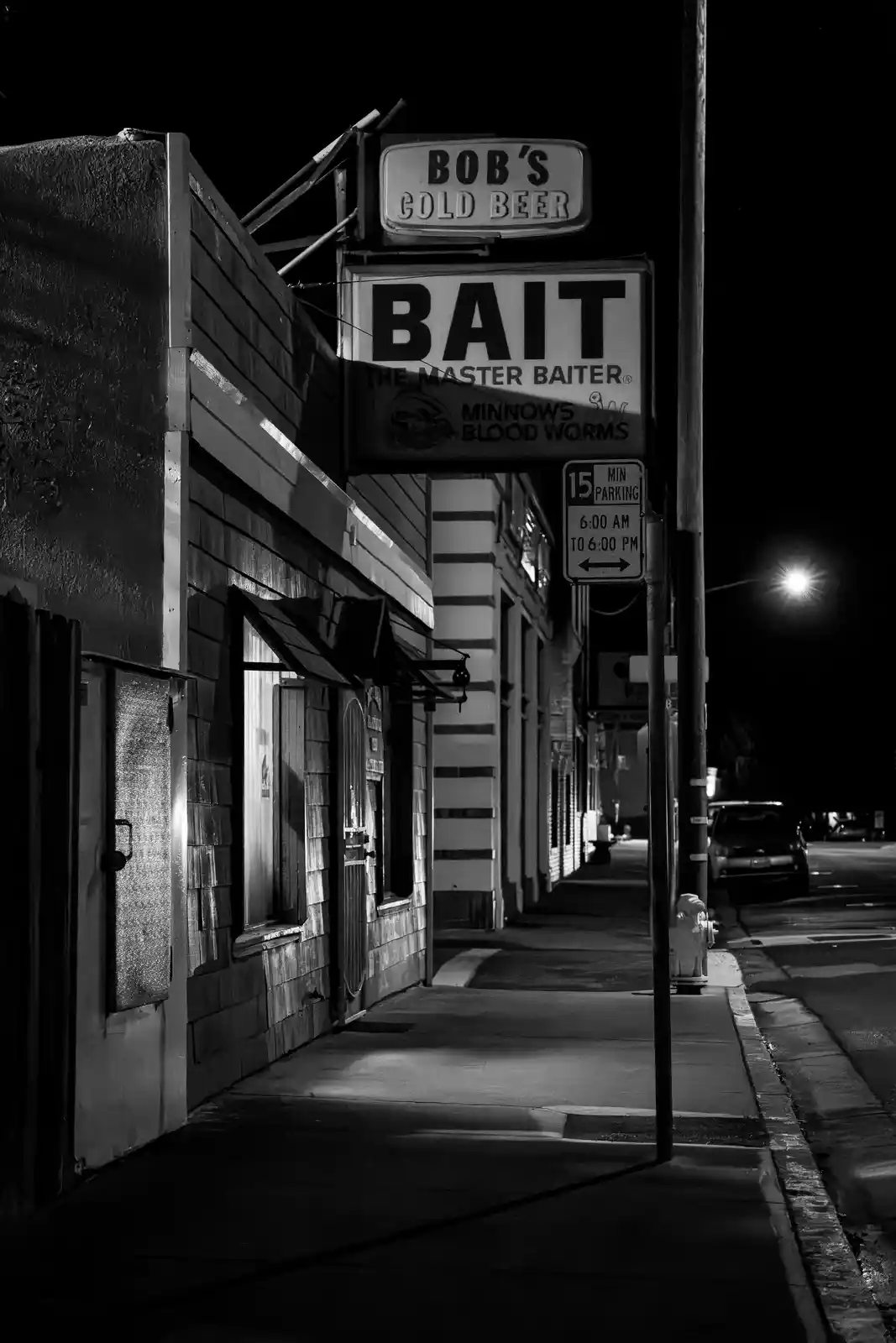 A black-and-white nighttime photo of a street corner with a store sign reading 'BOB'S COLD BEER' above a larger sign for…