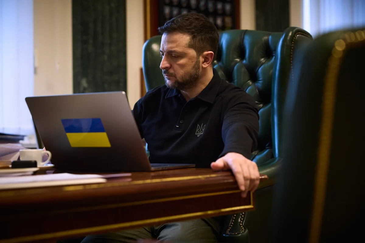 a man with a beard wearing a black polo shirt sits in a green leather chair at a wooden desk, looking intently at a laptop…