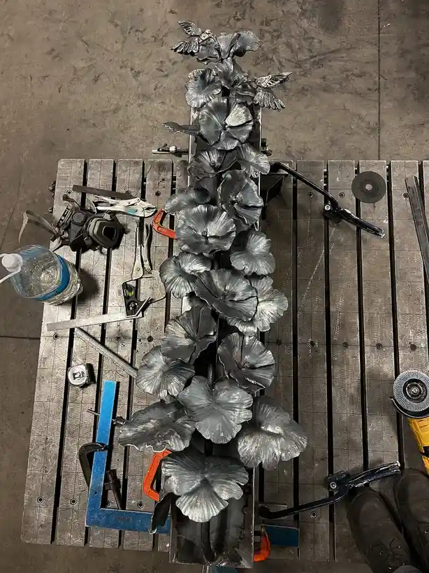 An overhead shot shows a metal sculpture of flowers and birds, clamped to a wooden workbench surrounded by tools like…