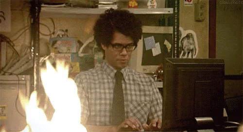 A man with curly hair and glasses sits at a desk in front of a computer, looking intensely focused as flames erupt from the…