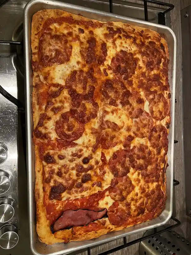 A rectangular pepperoni pizza on a metal baking sheet sits on a stovetop, with melted cheese, tomato sauce, and pepperoni…