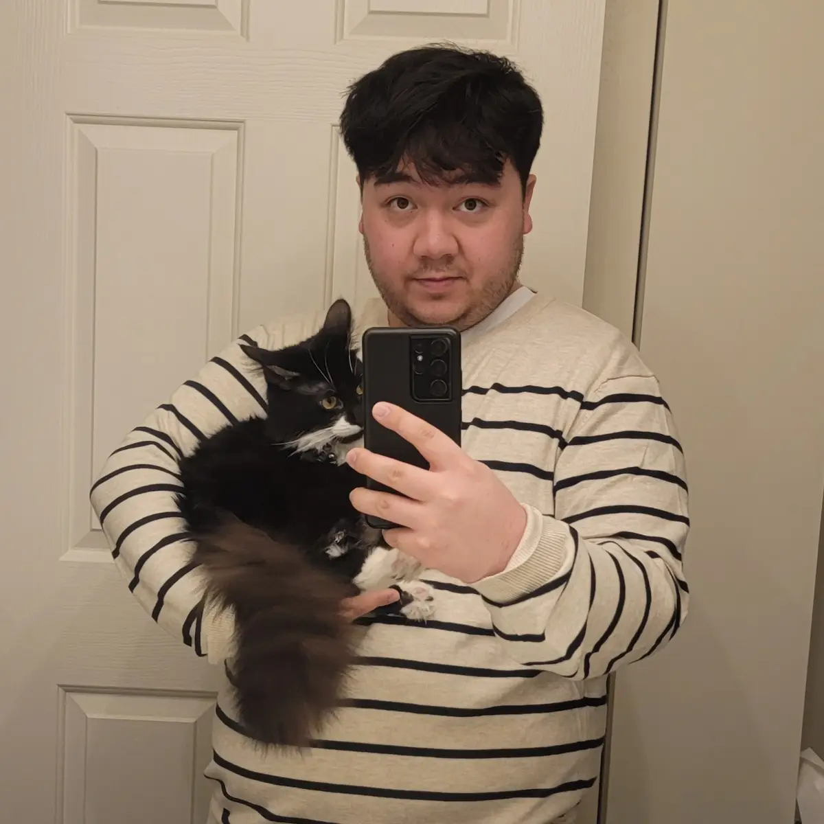 A person with dark hair and a striped shirt takes a mirror selfie while holding a fluffy black-and-white cat. The cat looks…