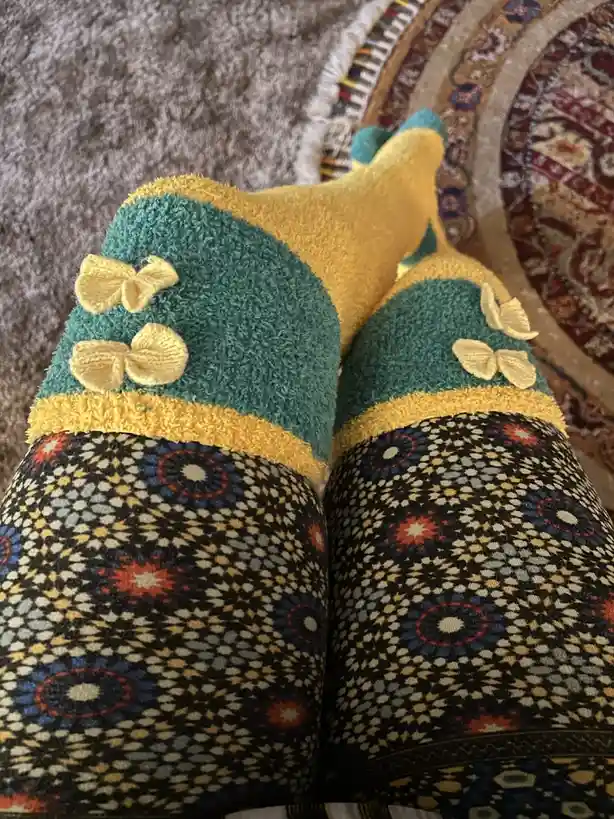 A close-up photo shows a person's feet wearing colorful, fuzzy socks with yellow bows on teal tops, paired with patterned…