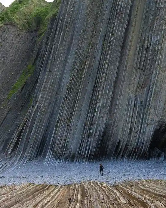 A person stands alone on a pebbled beach at the base of a massive, towering cliff composed of striking vertical basalt…