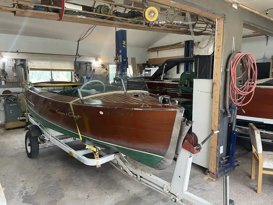 A vintage wooden Chris-Craft speedboat with hull number 20E9395 sits on a trailer inside a cluttered workshop filled with…