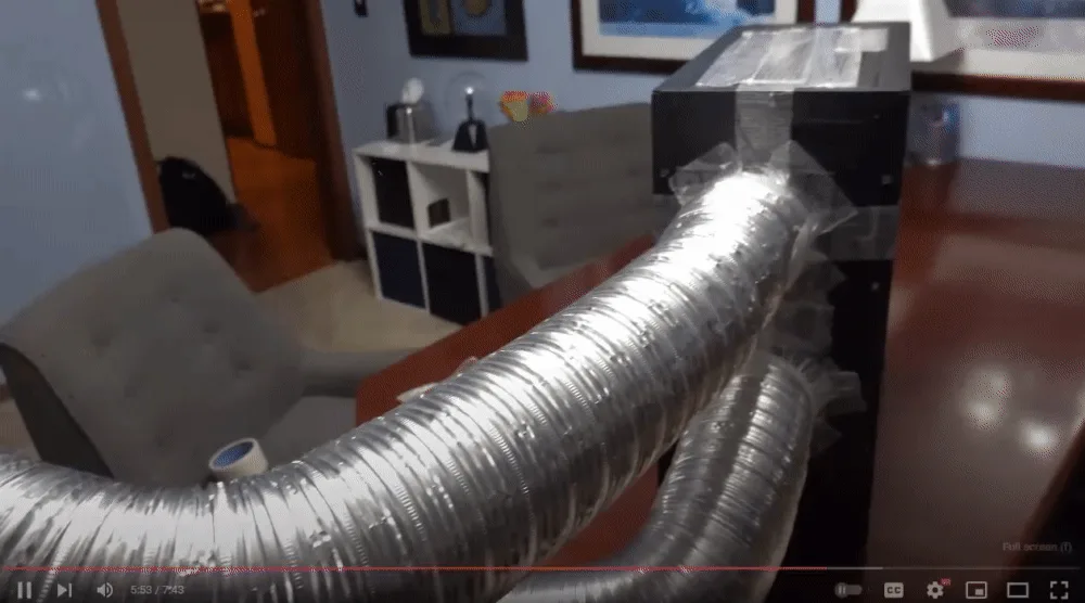 The image shows a split-screen video clip of a large, flexible, silver duct connected to a black box on a desk in an office…