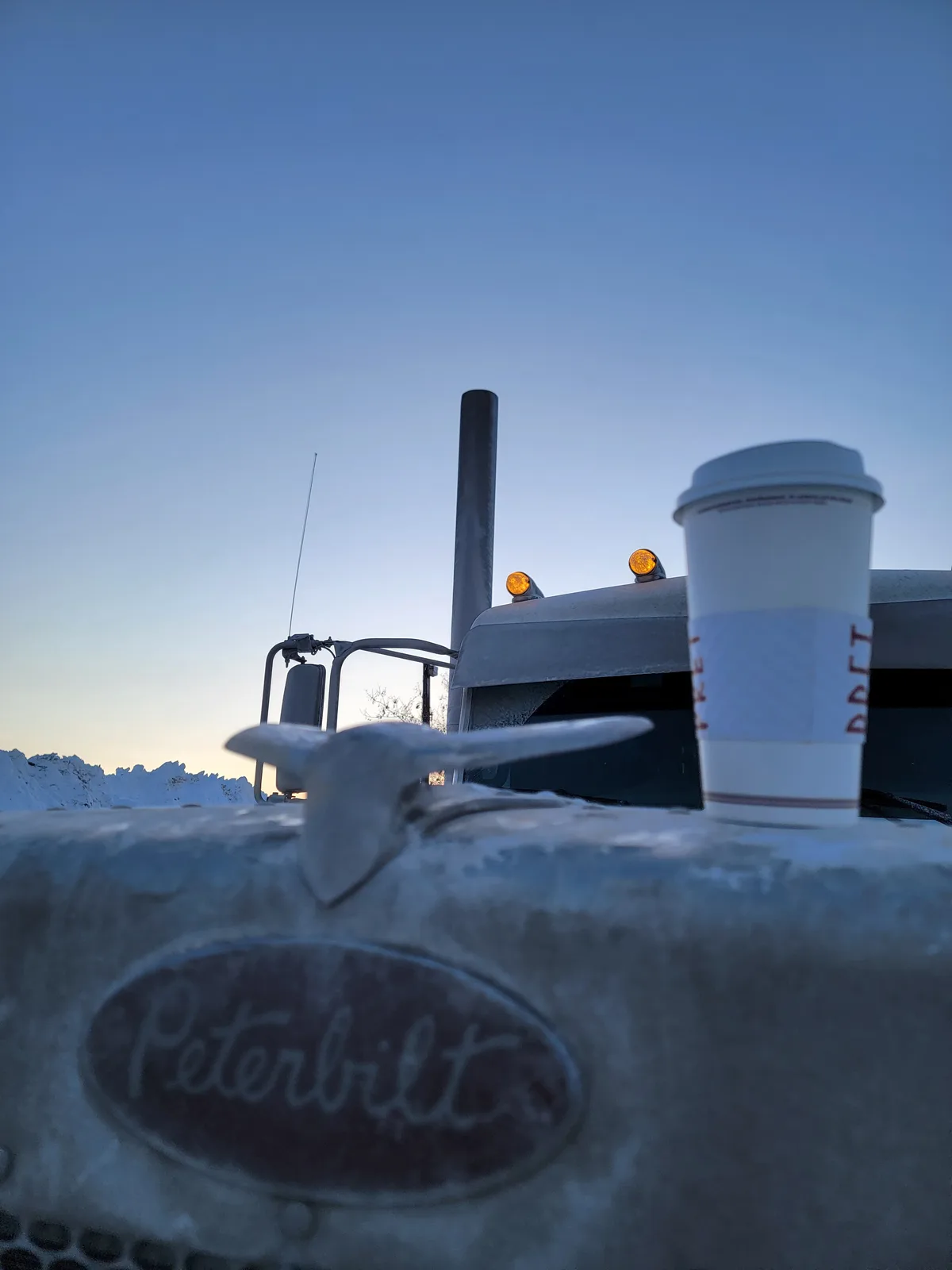 A white disposable coffee cup sits on the icy hood of a Peterbilt truck, with the truck's cab, smokestack, and orange…