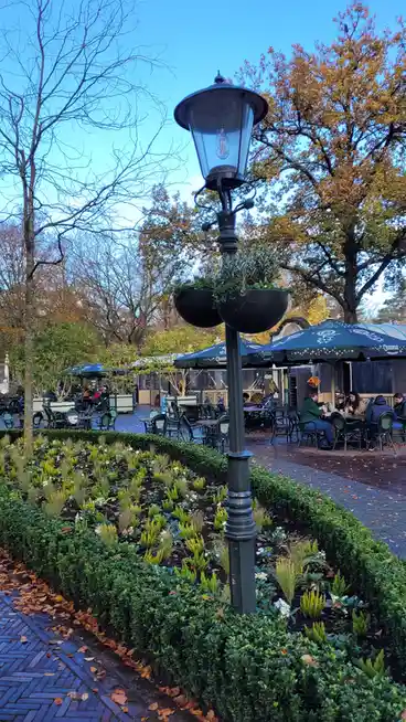A photo of an ornate black lamppost with hanging planters, surrounded by manicured hedges and autumn leaves, in an outdoor…