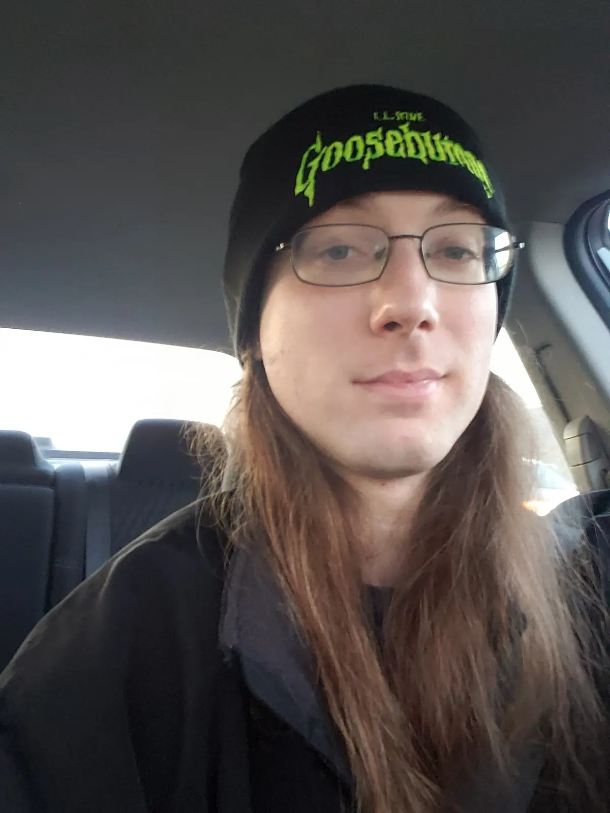 A person with long brown hair and glasses sits in a car wearing a black beanie with bright green 'Goosebumps' text. The…