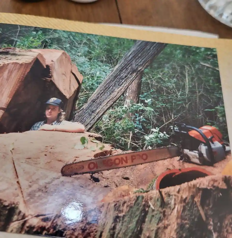 A printed photo shows a man with long hair and a baseball cap leaning against a large, freshly cut tree stump in a forest. A…