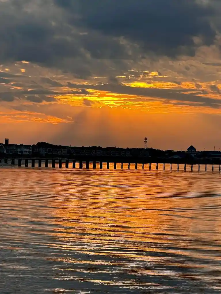 A serene sunset over water with a pier and houses silhouetted against a dramatic orange and yellow sky. The sun's rays…