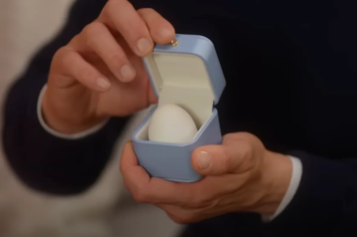 A person in a dark suit holds a small, light blue jewelry box with a white interior, opening it to reveal a plain white egg…