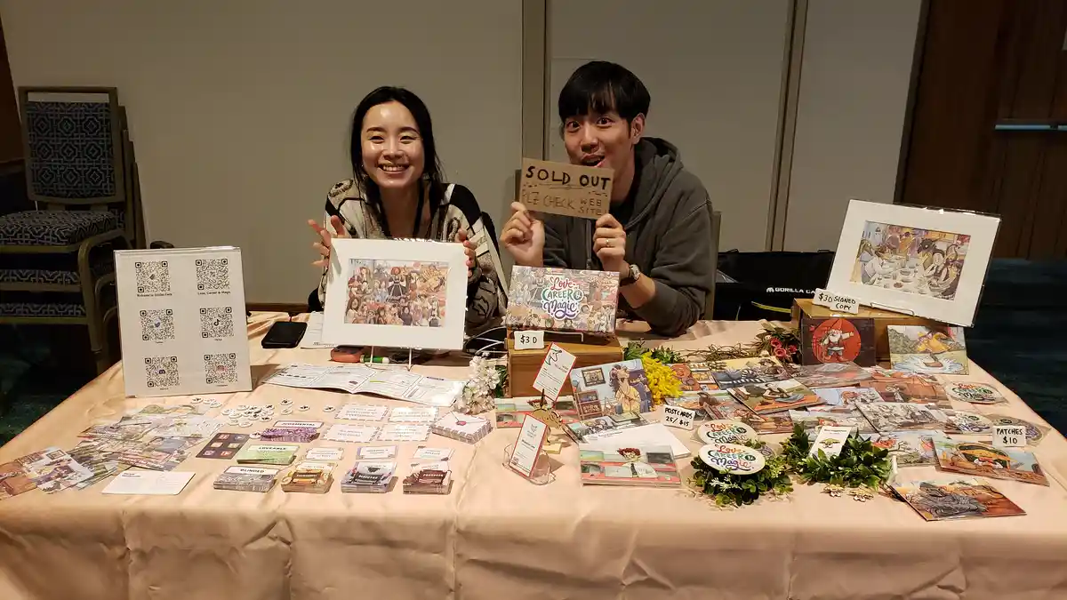 Two smiling people sit behind a table covered in art prints, cards, and merchandise for 'Love, Career & Magic'. The man on…