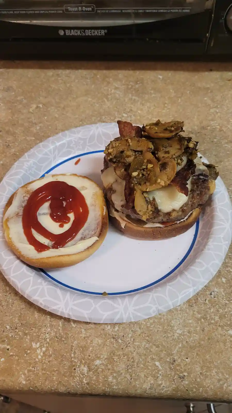 A homemade burger sits on a paper plate with its top bun separated, revealing a beef patty, melted cheese, bacon, and…