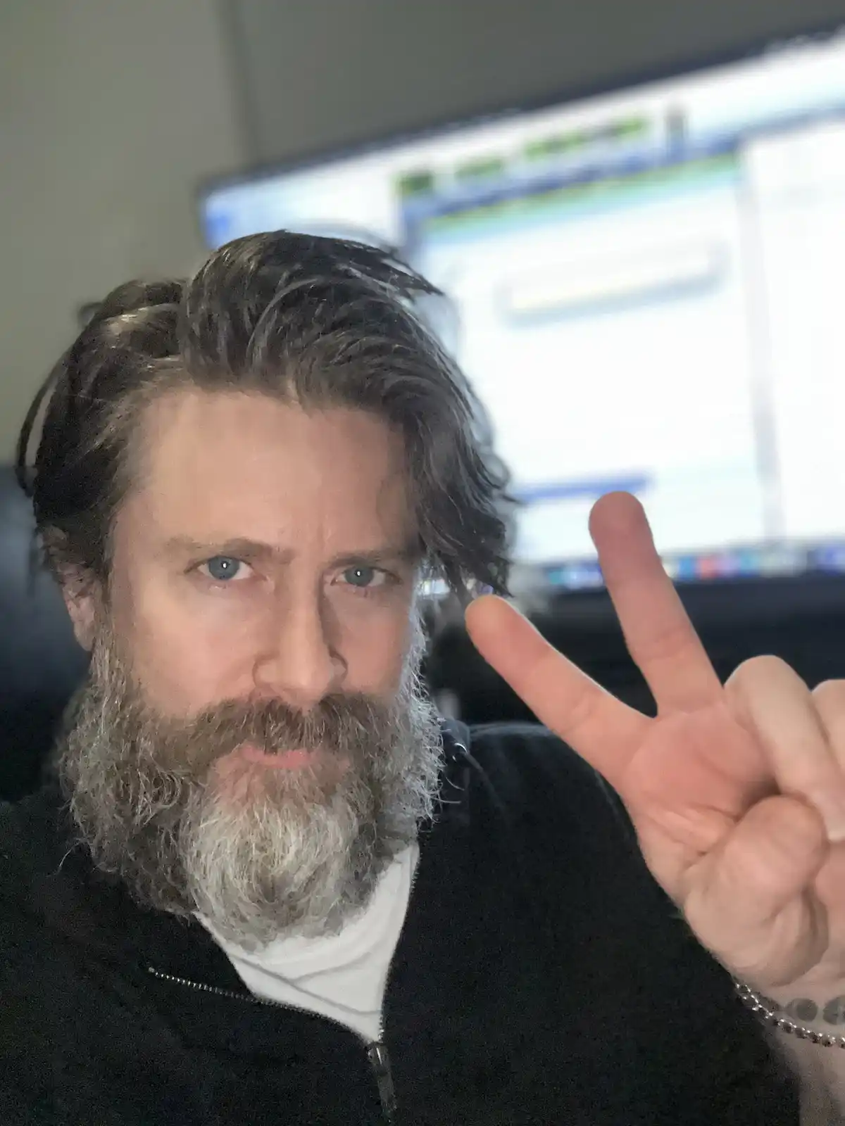 A man with a graying beard and styled hair makes a peace sign with his hand while looking directly at the camera, with…