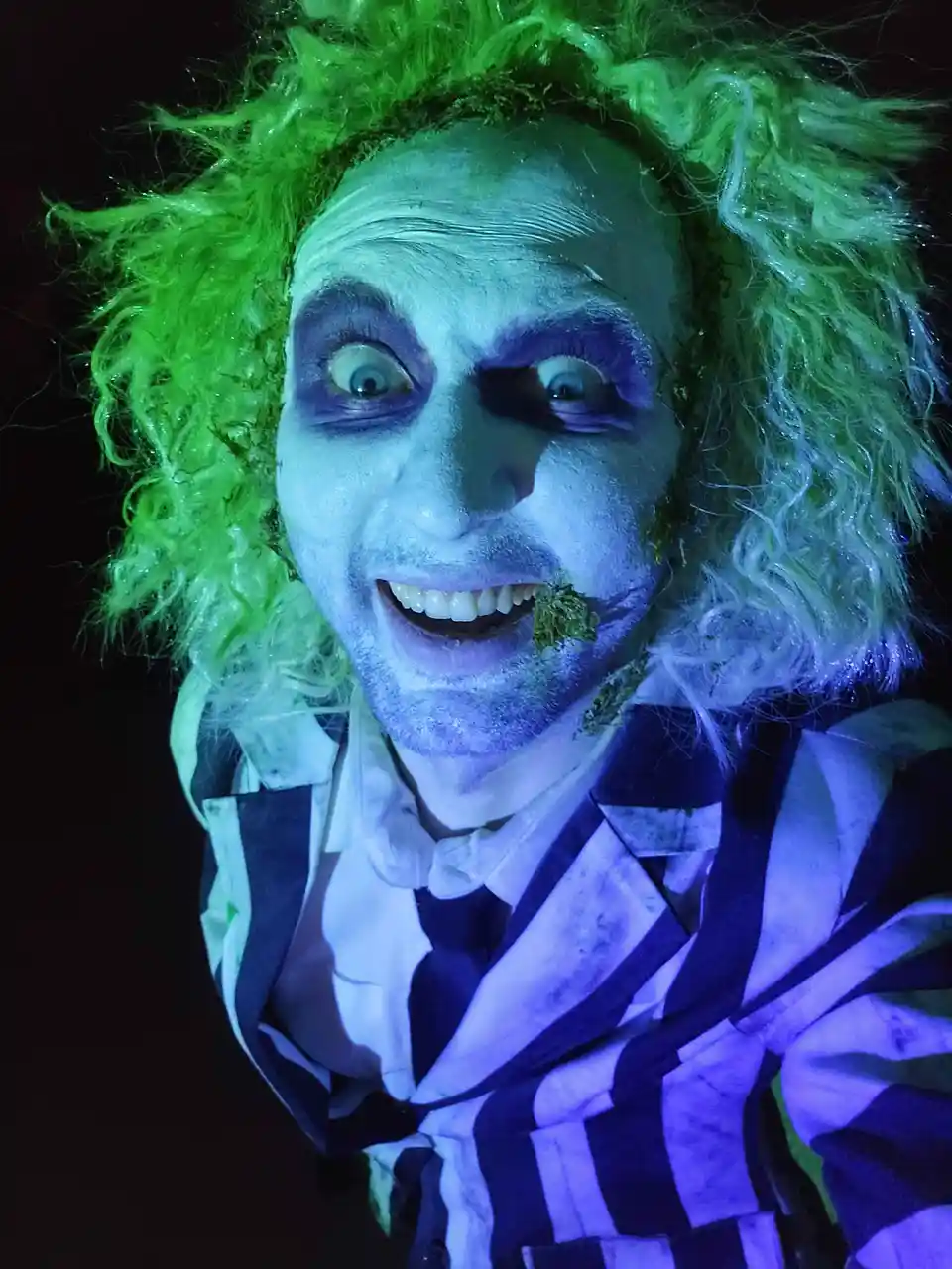 A person dressed as Beetlejuice from the movie, with bright green curly hair, pale face paint, purple eye makeup, and a…