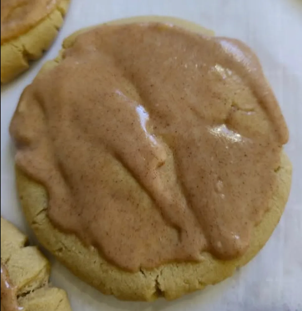 A close-up photo shows a round, golden-brown cookie generously covered with a thick, glossy, light-brown frosting speckled…