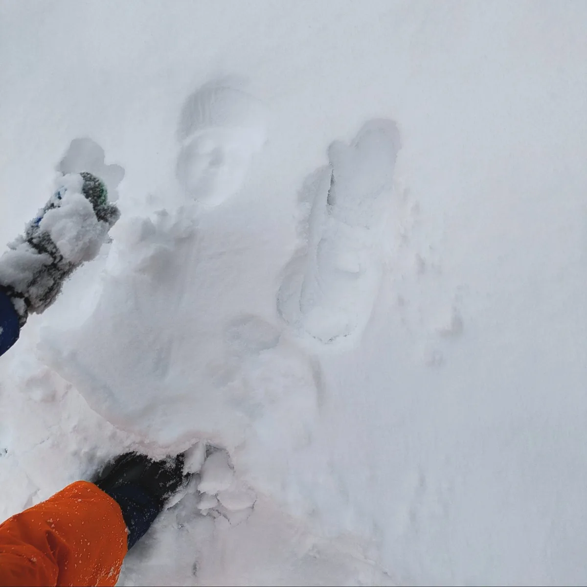 A person wearing an orange jacket and black gloves is partially buried in snow, with their gloved hands and one foot…