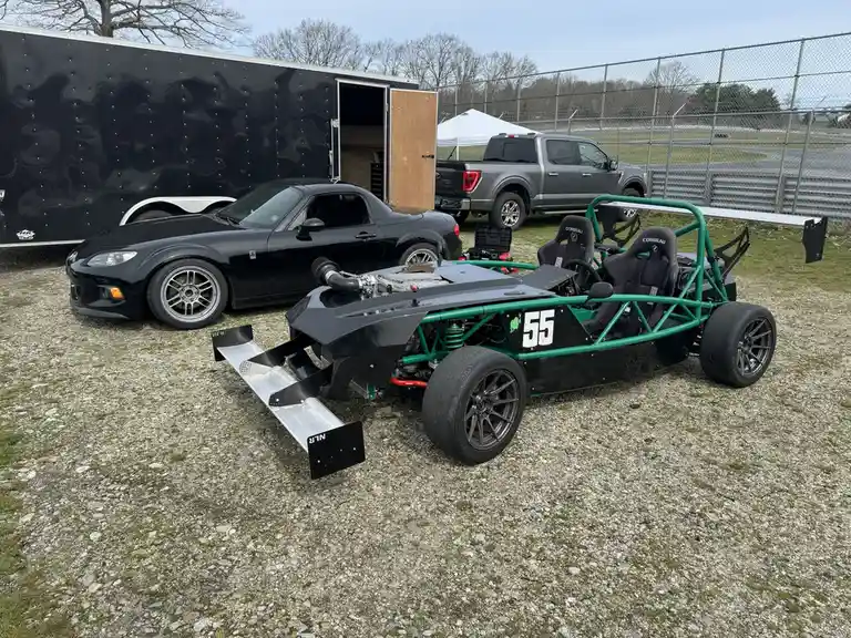 A black Mazda Miata is parked next to a green and black open-wheel race car with the number 55 on its side, both sitting on…