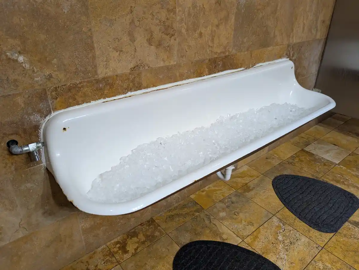 A white urinal mounted on a brown tiled wall is filled with crushed ice, with black foot mats on the tiled floor nearby. The…