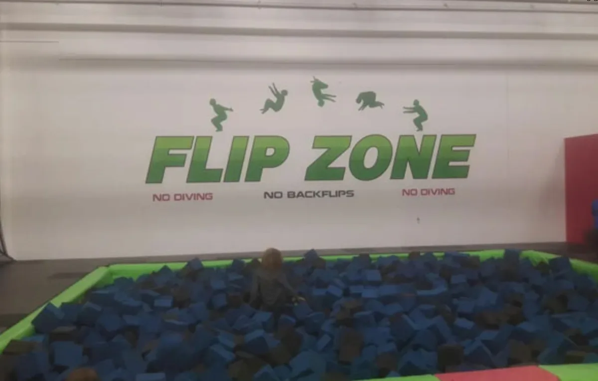 A person is partially submerged in a foam pit labeled 'FLIP ZONE' with safety warnings 'NO DIVING' and 'NO BACKFLIPS'…