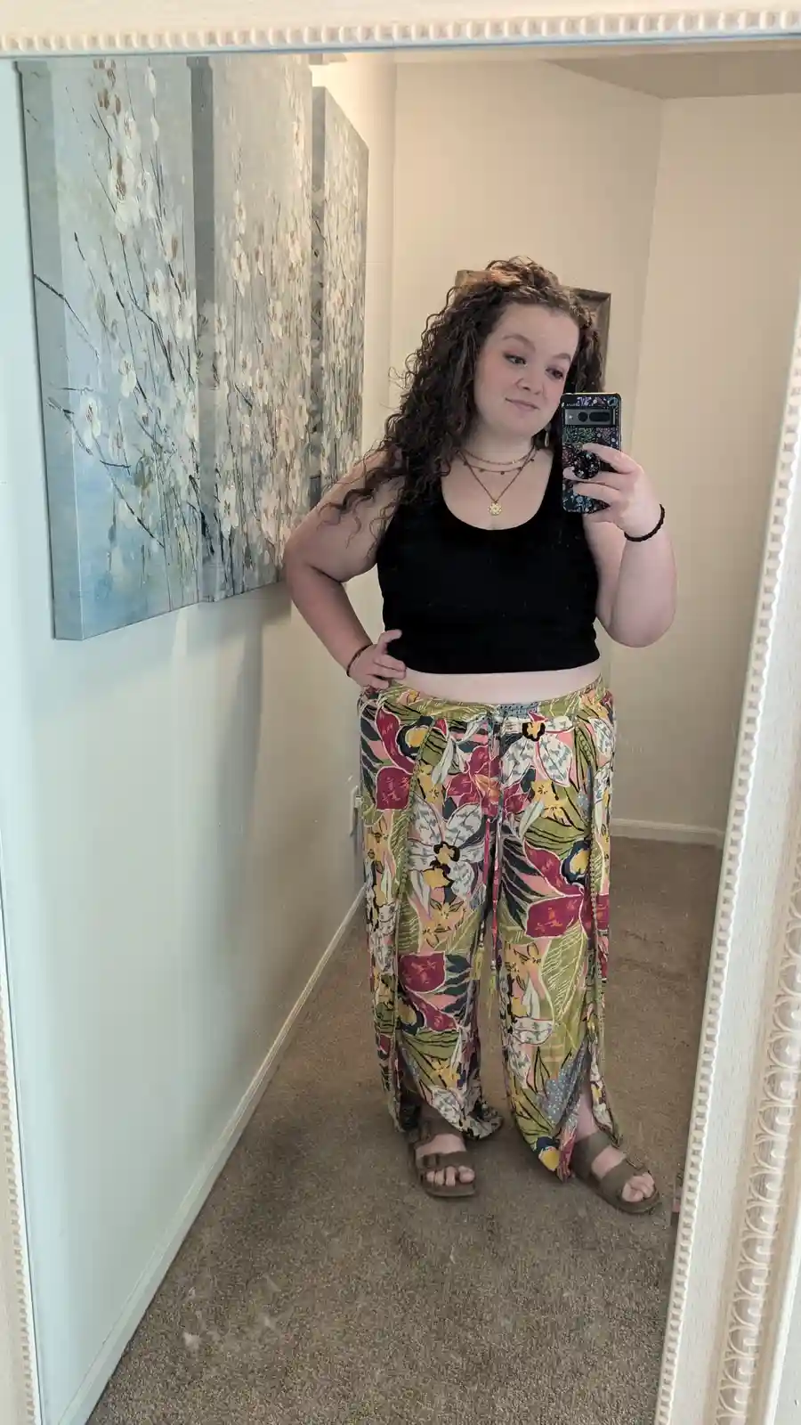 A person with curly hair takes a mirror selfie in a hallway, wearing a black crop top and colorful floral pants, holding a…