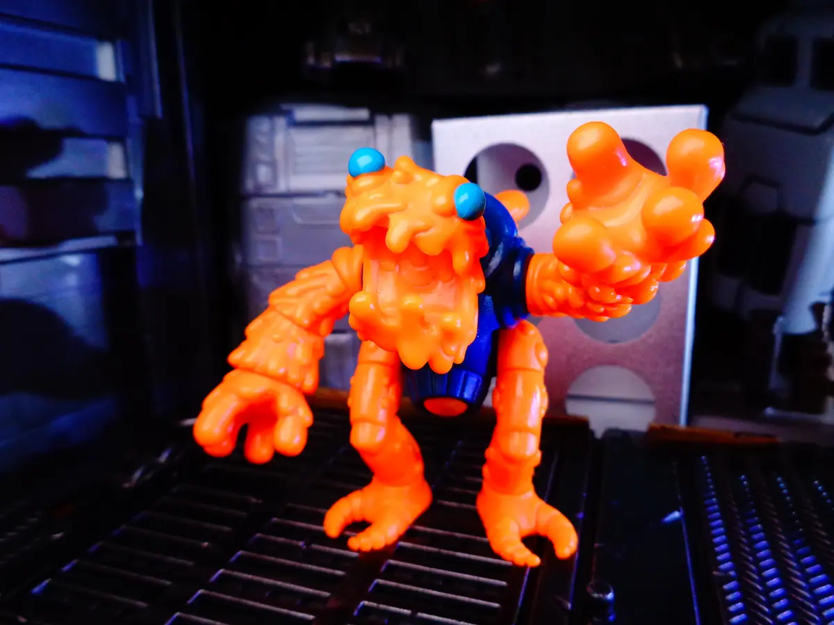 An orange, gooey, monster-like plastic toy figure with blue eyes stands on a black printer tray, surrounded by printer…