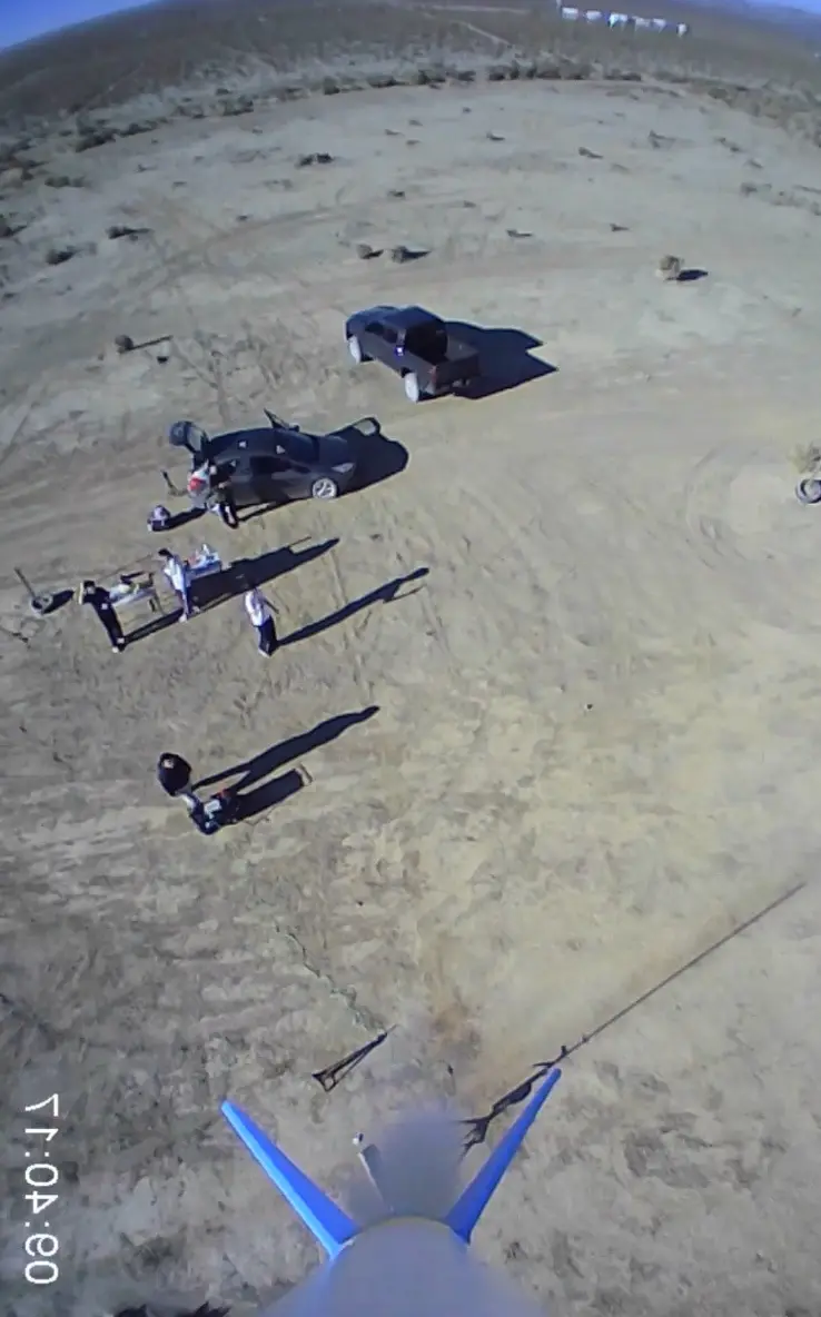 An aerial view from a drone or aircraft shows a desert scene with two cars and several people on the ground. One car is a…