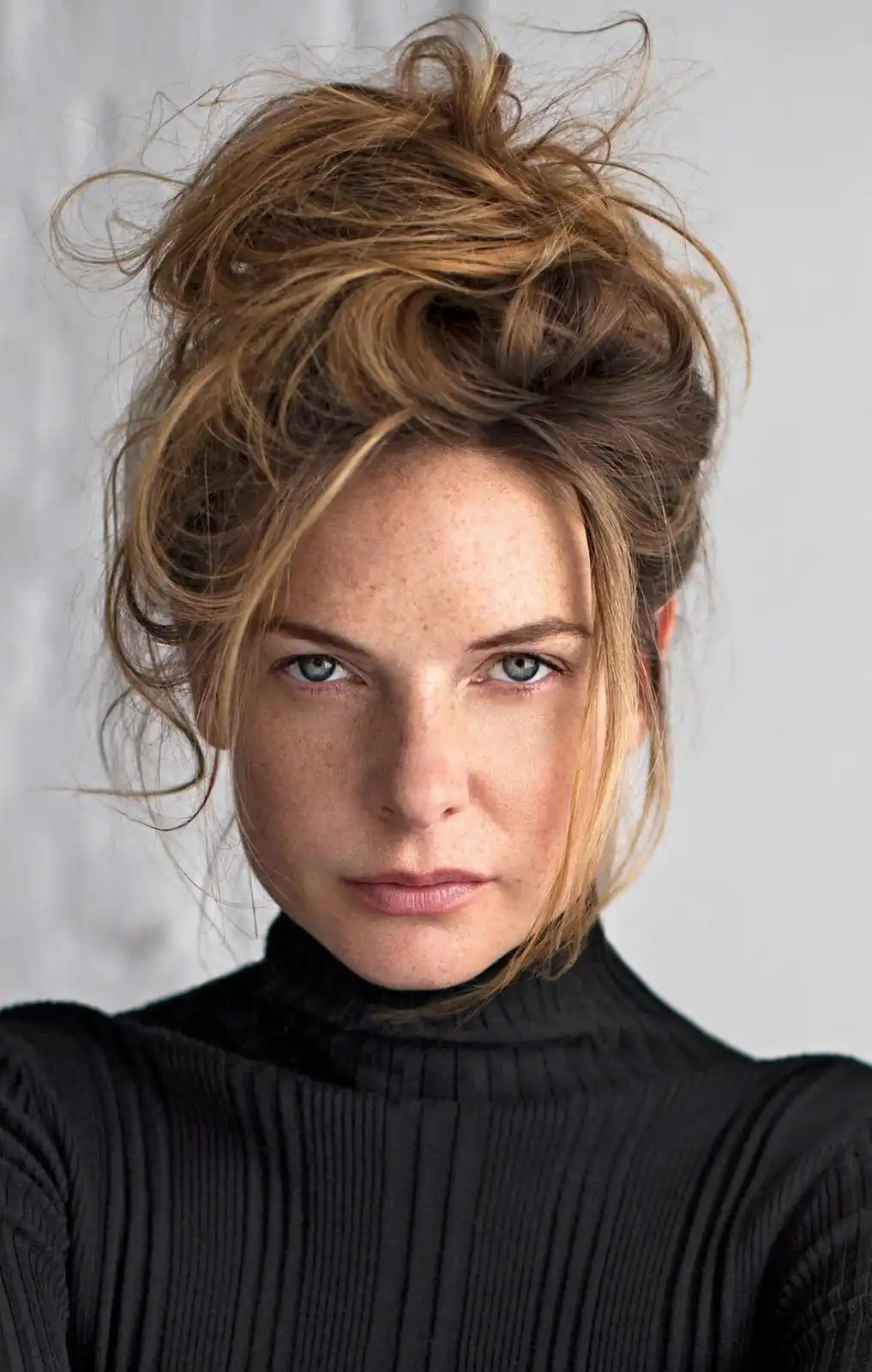 A woman with freckles, blue eyes, and messy brown hair styled in a bun looks directly at the camera with a neutral…