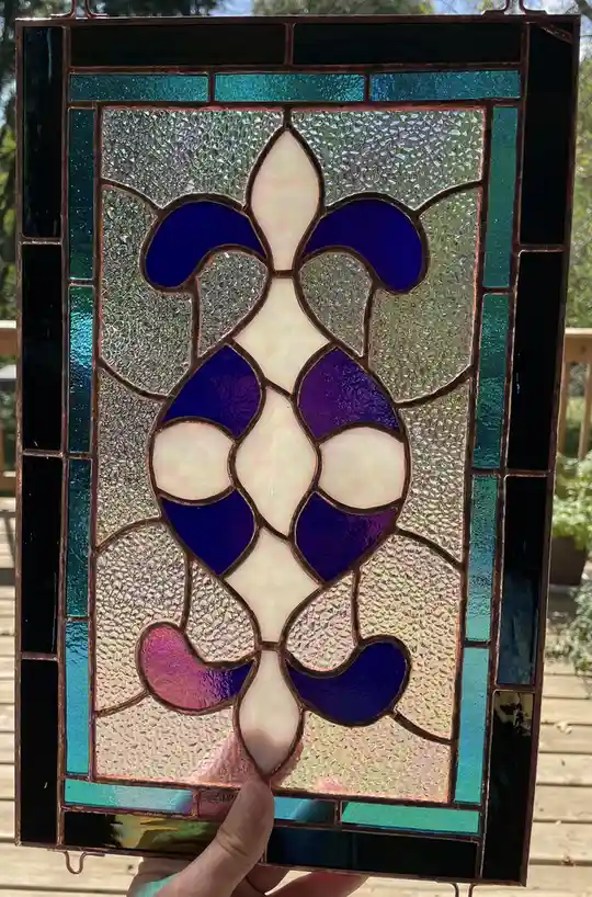 A hand holds up a rectangular stained glass panel featuring a central fleur-de-lis design made of blue, purple, and…