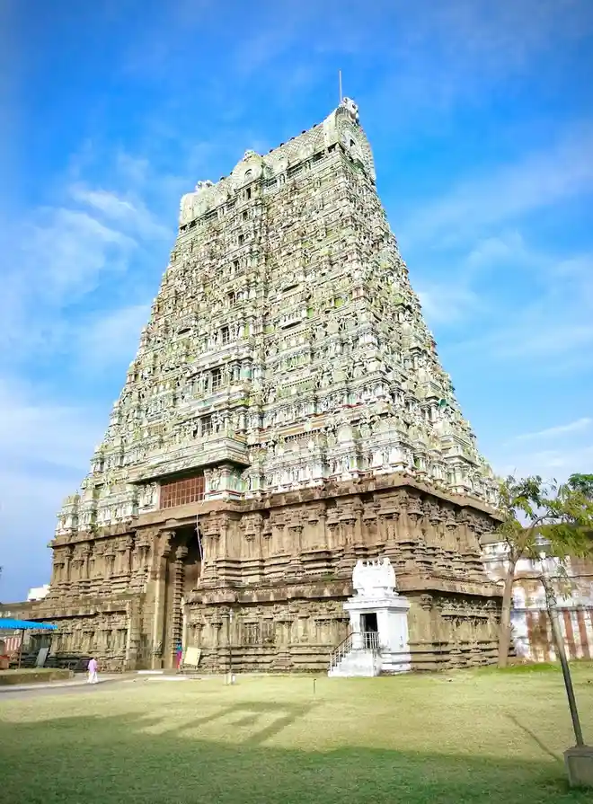 A towering, intricately carved Hindu temple with a steep, multi-tiered spire rises against a bright blue sky with wispy…