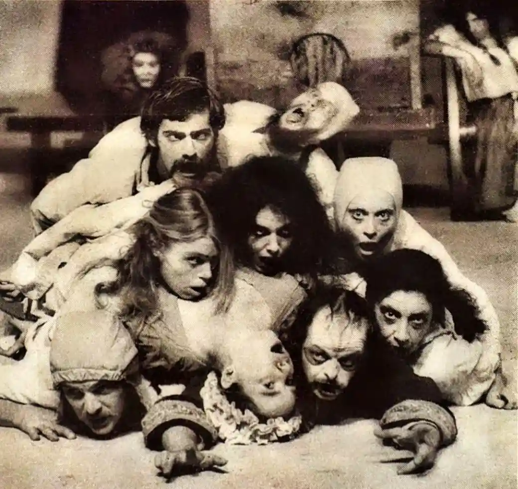 A black-and-white photo shows a group of actors in theatrical makeup and costumes, piled together in a chaotic, dramatic…