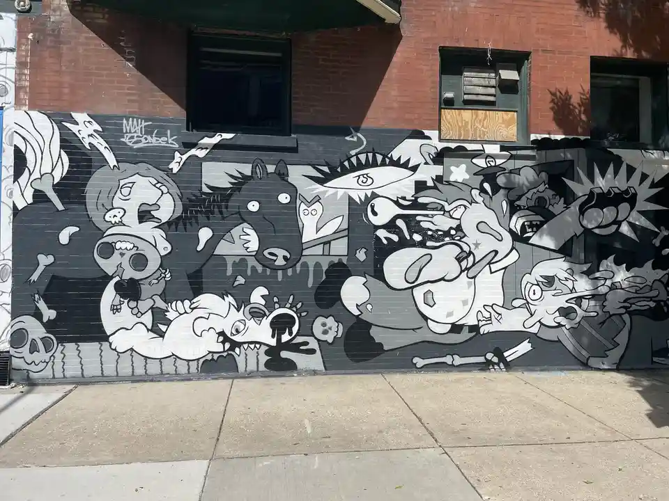 A black-and-white graffiti mural on a brick wall depicts a chaotic scene with cartoonish, exaggerated characters including a…